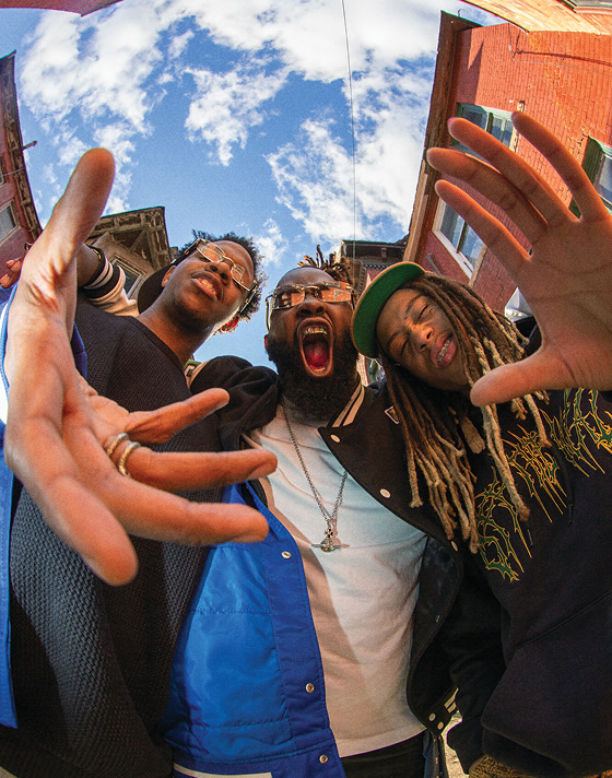 Hip-Hop/Rap trio, Hi-Tech, are making funny faces in front of a building, smiling and posing playfully for the camera.