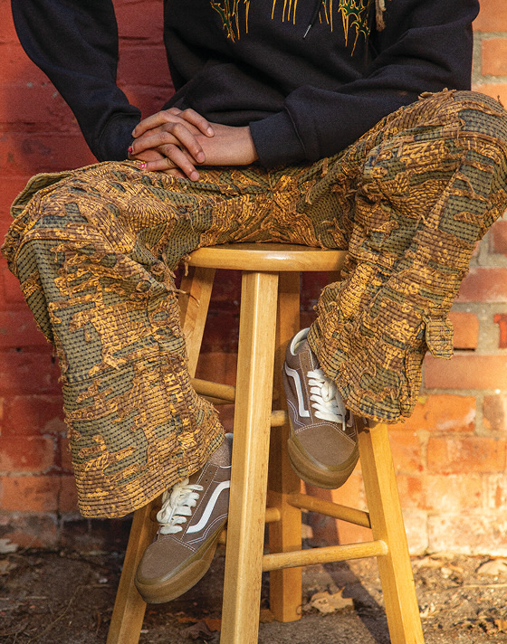 A person sits on a wooden stool behind a brick wall. Wearing dark pants, a black hoodie and Sepia Brown Premium Old Skool shoes.
