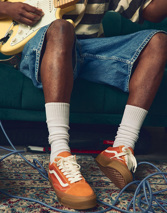 Close up of a person sitting on a couch, playing guitar, showcasing Marmalade Orange Premium Old Skool shoes with white socks.