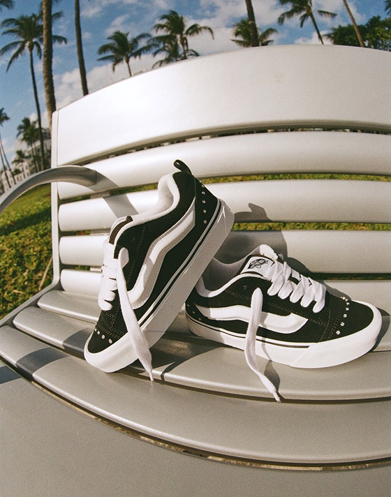 Close up of Rhinestone Black Knu Skool shoes resting on a bench. Behind them, a vibrant blue sky and palm trees.