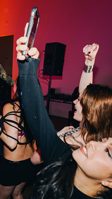 People dancing and taking selfies in a lively party setting with colorful, moody lighting.