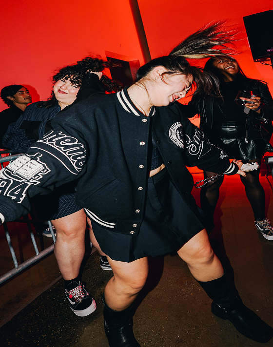 People dancing energetically at a party with red lighting. A woman wearing a varsity jacket is in focus, while others in view are wearing black Old Skools.