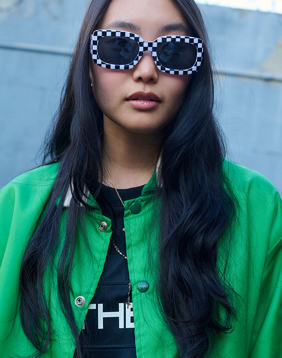 Person wearing checkered sunglasses, the Stevies Sports Jersey, and a green jacket, standing outdoors.