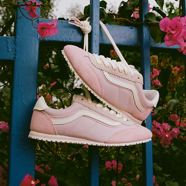 A pair of Sepia Rose Super Lowpro shoes. Its shoe strings are tied against a blue fence, surrounded by pink flowers. 