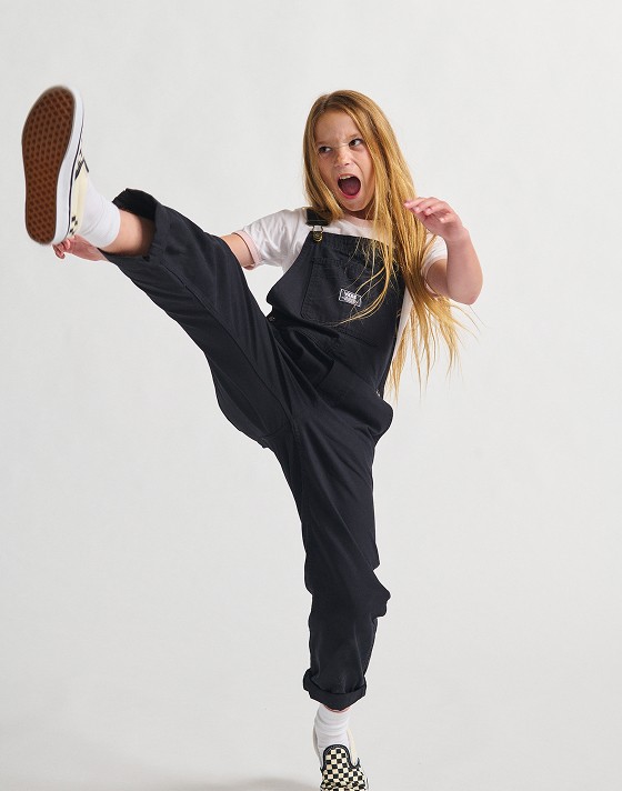 A girl with long blonde hair kicking the air wearing black overalls. 