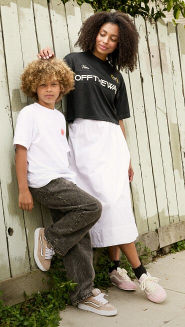 A woman and a child lean against a painted beige fence. The woman is wearing a Vans jersey, a white skirt, and Super Lowpro sneakers in Sepia Rose Pink. The child is wearing a white t-shirt, black jeans, and Hylane sneakers in Warm Taupe Brown.
