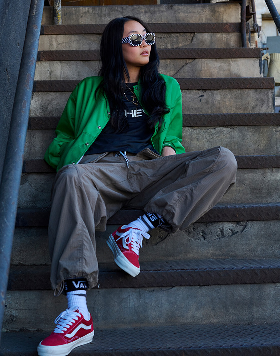 A woman sits on a set of concrete stairs looking off into the distance. She wears checkerboard sunglasses, a green jacket, the Stevies Sports Jersey, olive pants, and Premium Old Skool Suede sneakers in red.