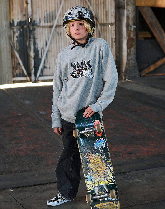 A child with long blonde hair stands in a warehouse, wearing a helmet and holding a skateboard upright. He wears a grey graphic crewneck, baggy black jeans, and Classic Slip-On sneakers in Checkerboard.