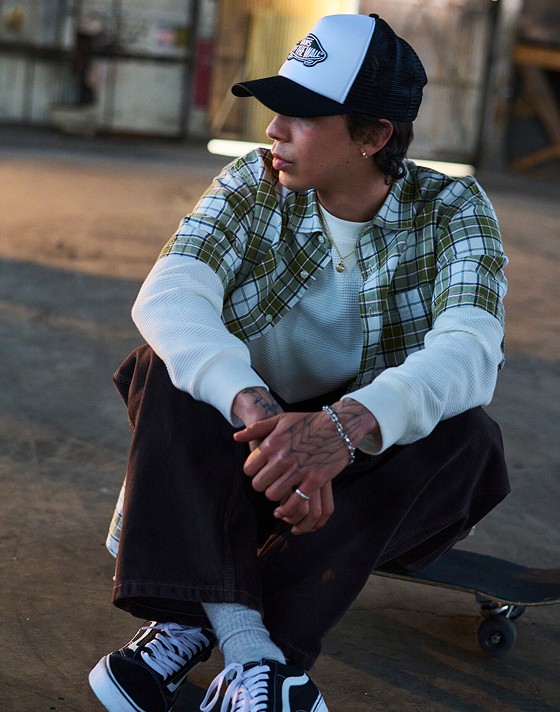 A person sits on a skateboard cross-legged inside a warehouse. They are wearing a black and white Vans trucker hat, a short sleeve green plaid shirt with a white thermal underneath, brown jeans, and Old Skool sneakers in black.