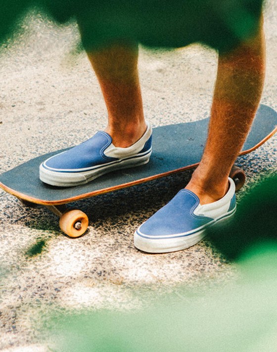 A knee-down shot of a person standing on concrete with one foot on a cruiser. Foliage frames the shot, and the person wears Slip-On Reissue sneakers in Thalia Blue.