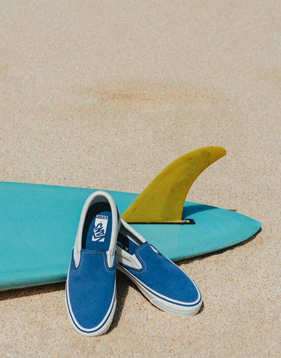A pair of Slip-On Reissue sneakers in Thalia Blue sit on a multicolored surfboard lying in the sand.