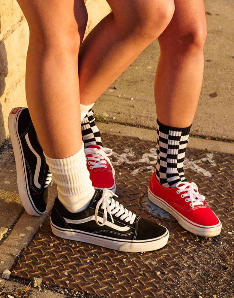 An image of two people from the calf down standing on a sidewalk. One person is wearing white socks and black and white Old Skool shoes. The other person is wearing black and white checkered socks and red and white Authentic shoes.