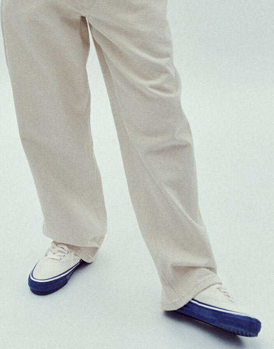 A detail waist-down shot of a person standing against a white studio background. They are wearing cream jeans and Premium Authentic 44 sneakers in Duck Canvas White & Blue.
