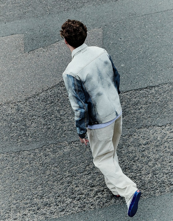 A Dutch angle shot of person walking away from the camera across a road. They are wearing a white and blue denim jacket, cream jeans, and Premium Authentic 44 sneakers in Duck Canvas White & Blue.