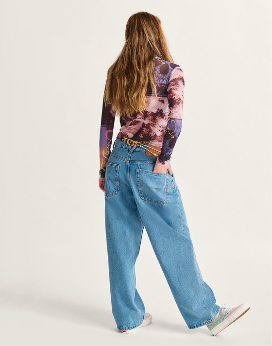 A person stands facing away from the camera on a white studio backdrop. They are wearing the Ludlow Mesh Long Sleeve, blue jeans, and Premium Authentic 44 sneakers in Checkerboard Sepia Rose.
