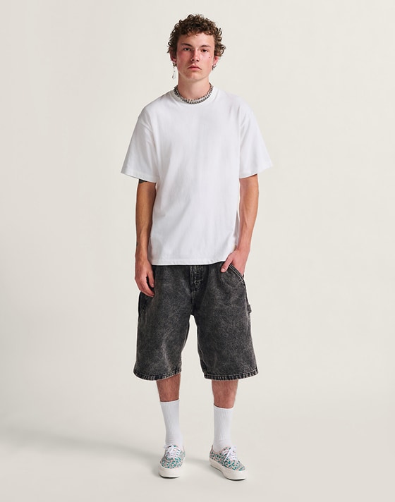 A person stands facing the camera on a white studio backdrop. They are wearing a white t-shirt, black denim shorts, and Premium Authentic 44 sneakers in Cheetah Pool Blue Multi.