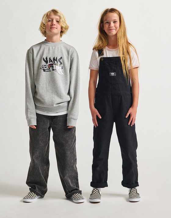 Two children stand facing the camera on a white studio backdrop. One is wearing a grey crewneck and black jeans, and the other wears a white and pink ringer t-shirt and black overalls. They both wear Classic Slip-On sneakers in checkerboard.