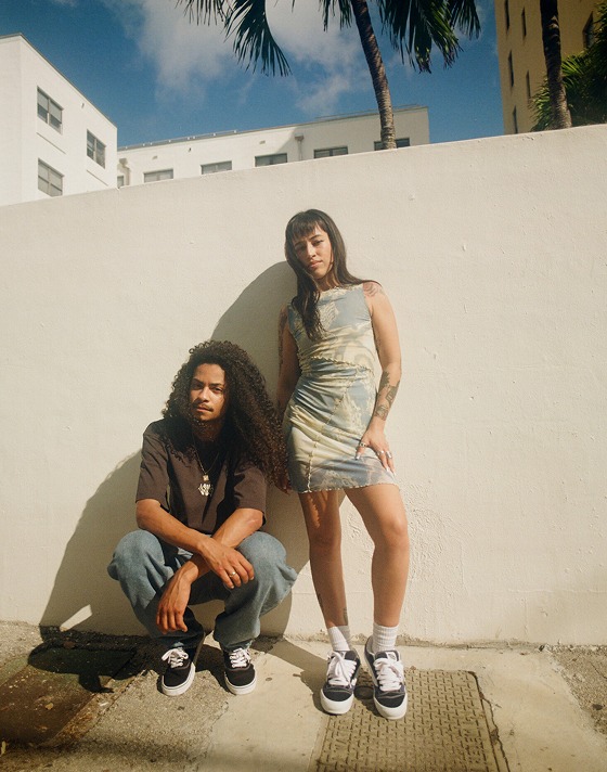 A man and woman pose next to a wall. The man squats and wears black Authentic shoes while the woman wears a skirt and black and white Knu Skool.