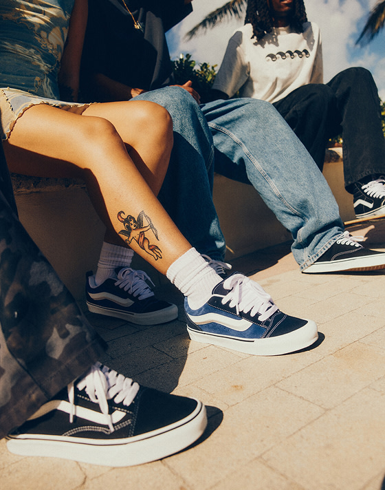 Close-up of young people sitting closely on a bench, wearing different Vans shoes, with the focus on a young woman sporting bold Knu Skool shoes.