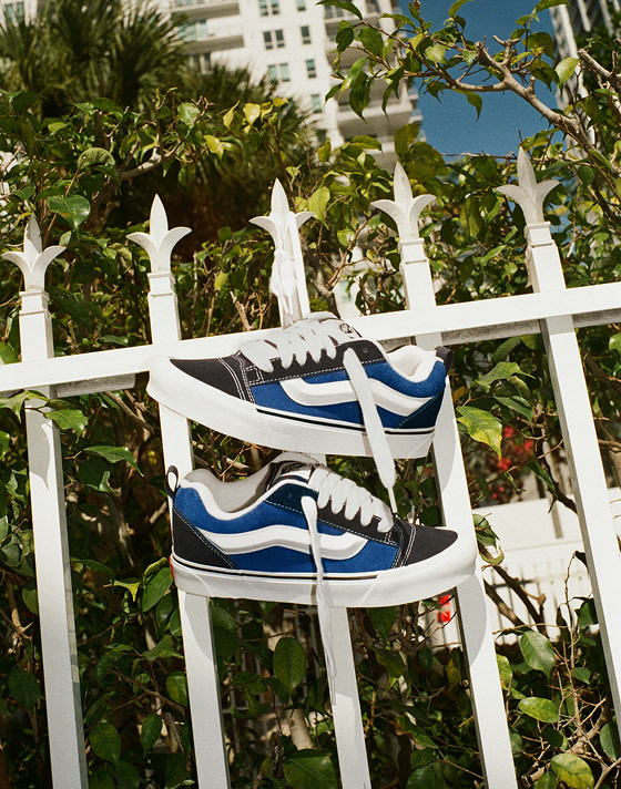 A pair of black and blue Knu Skool hang from a white pointed metal fence with their laces tied together with trees in the background.