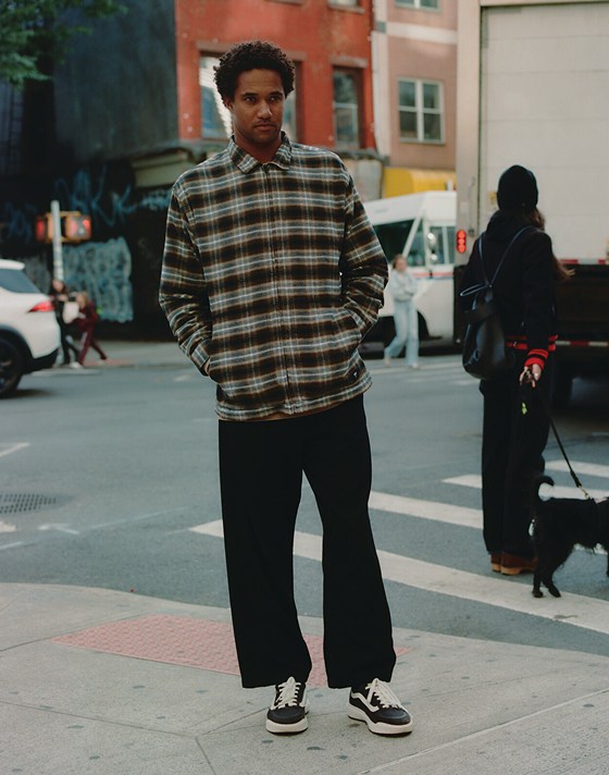 A man stands at a busy street corner wearing UltraRange 2.0 shoes, blue jeans, and a plaid long-sleeve jacket.