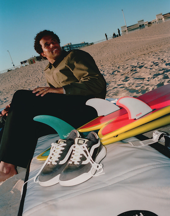 A young man at the beach prepares to surf on a sunny day, his UltraRange 2.0 shoes casually placed on his surfboard, in a candid, unposed moment.