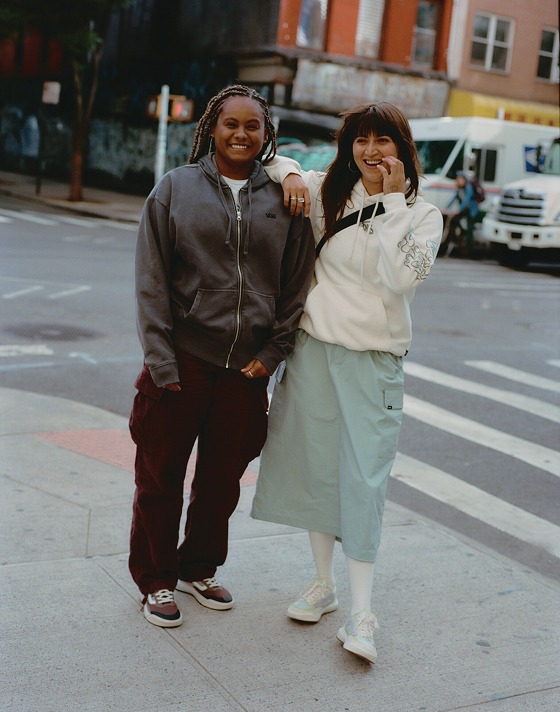 Two women pose on a street corner. One wears a Vans hoodie and red cargo pants while the other is in a white Vans hoodie and a skirt.