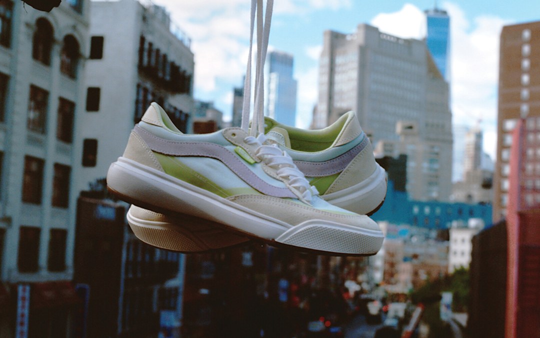 A pair of UltraRange 2.0 shoes dangle midair against a sunlit, slightly cloudy sky, with a blurred cityscape in the background adding depth and motion to the scene.