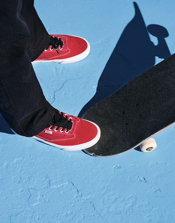 Close-up of a pair of red Curren shoes in a powder blue skate bowl with one foot on a skateboard's tail.