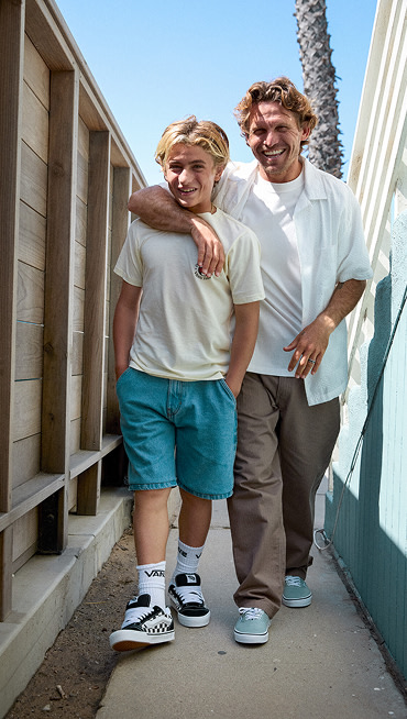 A father with his arm around his son walks down an alley. The father wears a white overshirt and Authentic while the son wears Checkerboard Knu Skool.