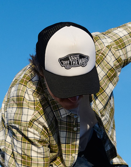 A close-up of a Vans "Off the Wall" trucker hat on a man wearing a flannel outside on a clear day.