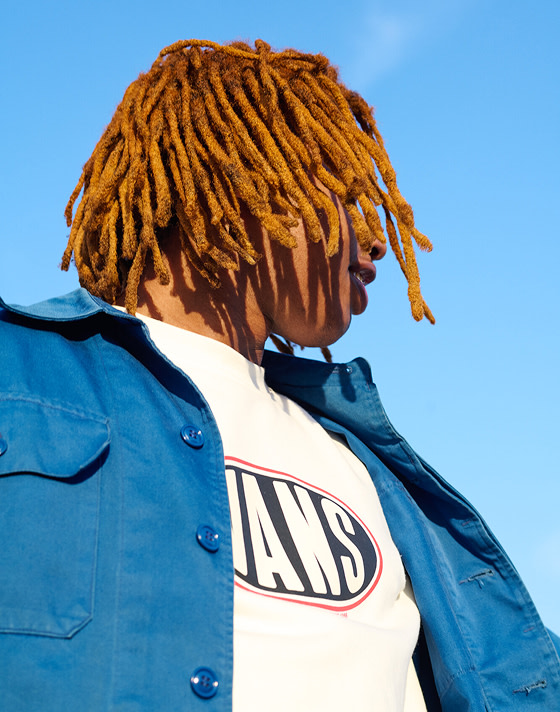 A man with gold-dyed dreads wearing a white Spray On Loose Crew Sweatshirt under a blue overshirt with the sky behind him.