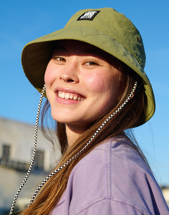 A woman smiling wearing a green Vans bucket hat with a blue sky behind her.