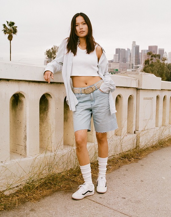 A woman leans up on a concrete guardrail in the Gillian Striped Button Up Shirt, Sirelle Cut-Off Jorts, and white Super Lowpro with a city behind her.