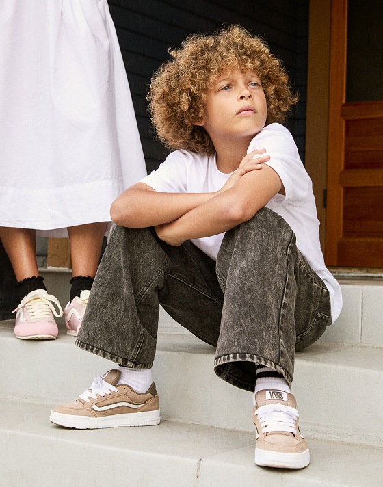 A boy sits on some steps wearing a white t-shirt, black jeans, and brown Hylane shoes.