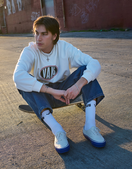 A man sits on his skateboard in a parking lot wearing a Spray On Loose Crew Sweatshirt and Premium Authentic during golden hour.