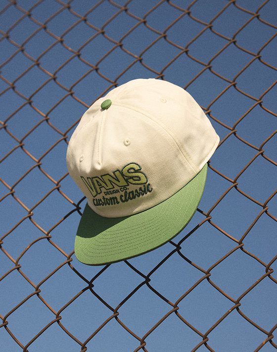 A white and fairway green Vans Design Co. Low Hat hanging from a cyclone fence.