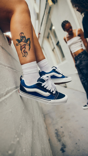 Somebody with an angel tattoo on their calf sits on a white brick wall wearing blue and black Knu Skool.