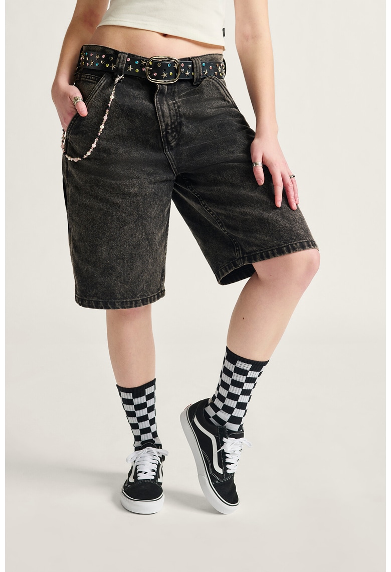 A waist-down shot of a woman wearing black denim shorts, Checkerboard socks, and black and white Old Skool.