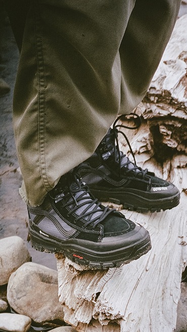 A knee-down shot of a man on a driftwood log wearing black Crosspath XC GORE-TEX on a wintry beach.