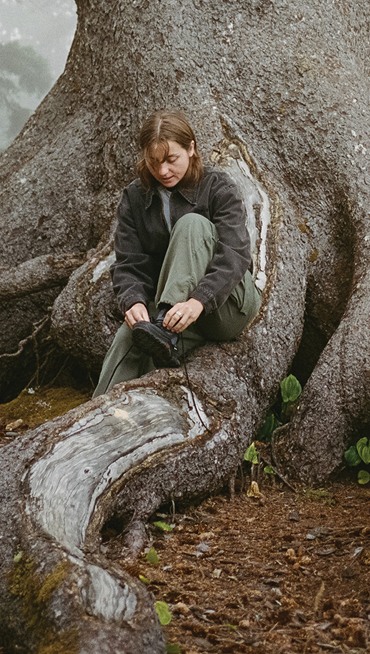 Wide shot of Vans Snowboarder Savannah Shinkse sitting on a root of a big tree lacing up a pair of Sk8-Hi GORE-TEX Insulated Shoe in black.