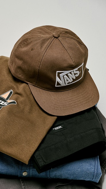 Close up of the Vintage Cocoa Brown Vans Formula Snapback Hat sitting on top of a pile of clothes in a grey photography studio.