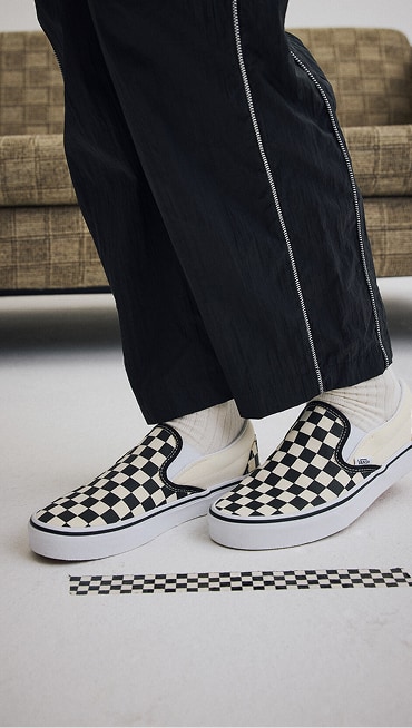 Close-up of a woman wearing black and white Checkerboard Classic Slip-On in the black Nyla tracksuit in a photography studio.