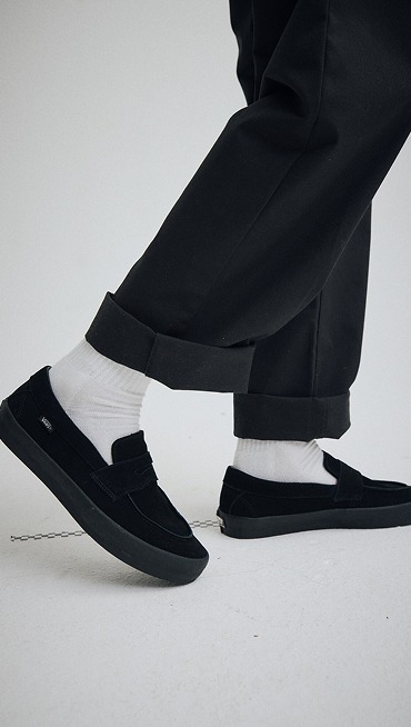 Knee down shot of someone wearing black pants and Black Suede Skate Loafers in a grey photography studio.