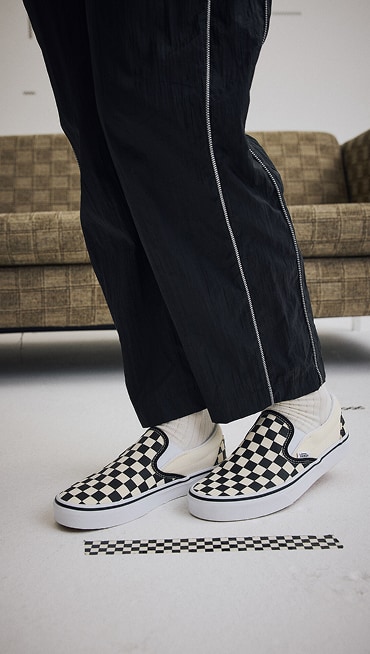 ​​Knee down shot of someone wearing the Nyla Track Pant and a pair of the black and white Classic Slip-On Checkerboard Shoe in a grey studio.