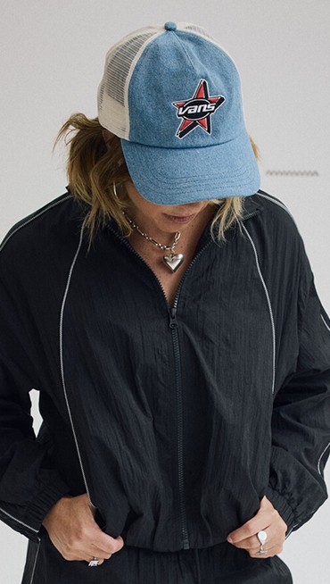 Medium shot of a woman wearing a Black Nyla Tracksuit and a Marked Jockey Trucker Hat in a grey photography studio.