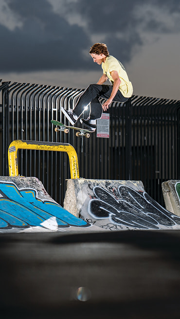 Wide shot of Curren Caples mid air skating off a graffiti covered cement ramp onto a worn down yellow rail.