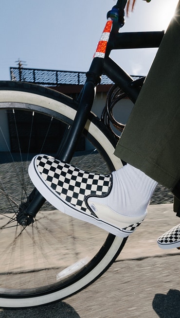Close-up of someone biking outside wearing white and black Checkerboard Classic Slip-On shoes.