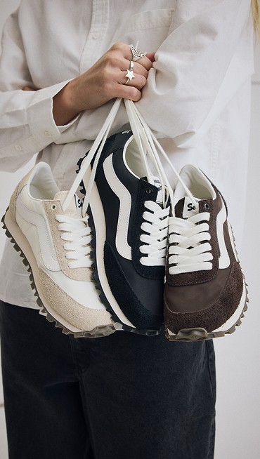 A young woman holding all three white, black, and brown colorways of the Premium Style 152 by their laces in a photography studio.