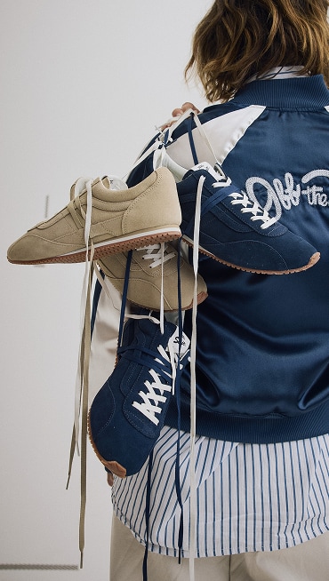 A young woman holding two pairs of Premium Super Lowpro Trainers by their laces over her shoulder in a photography studio.